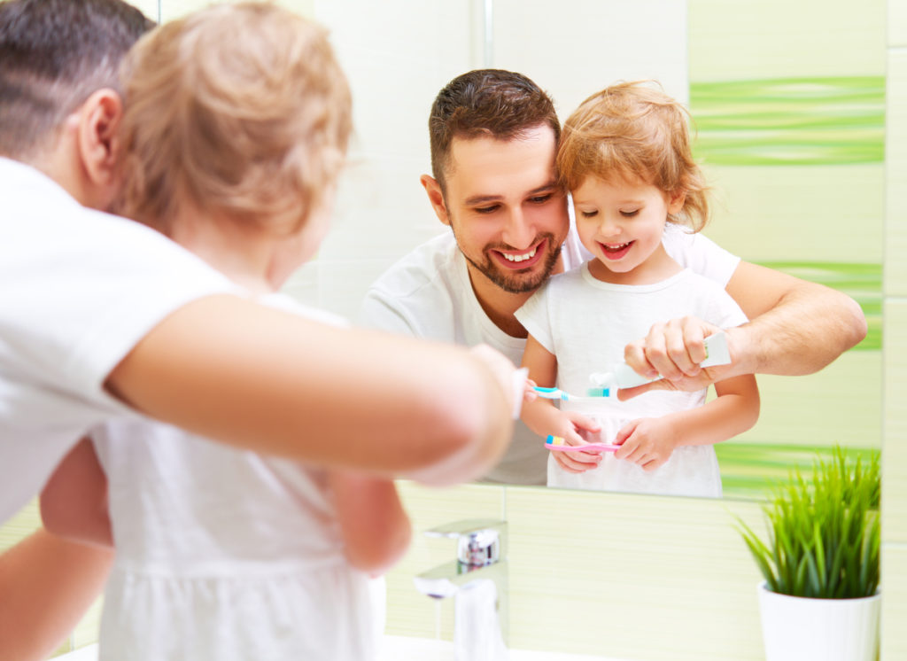 Niño cepillándose los dientes con su padre frente al espejo.